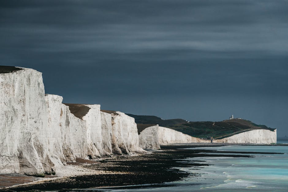 A panoramic view of the white chalk cliffs along the coastline, with some green grassy areas on top, under a cloudy sky. The cliffs extend into the distance, gradually shrinking in size, and a lighthouse is visible on a hilltop further along the coast. The shoreline features a dark pebble beach with wet sand, and the sea has gentle waves lapping at the shore. The scene depicts a natural landscape suitable for house removals and home relocation services, highlighting the importance of careful furniture transport and packing during the moving process, as organized by companies like Man with Van Seven Sisters.