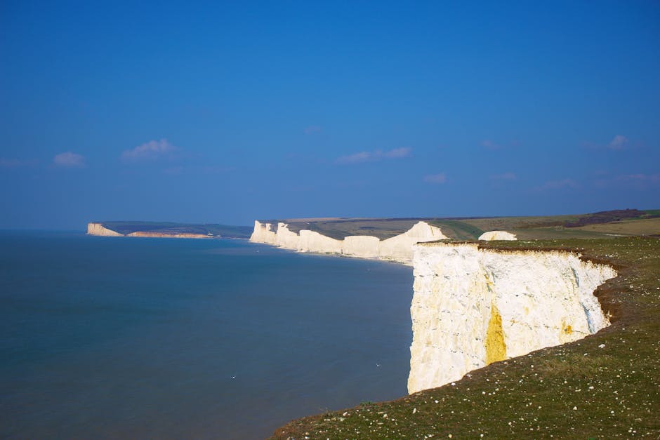 A panoramic view of the white chalk cliffs along the coastline, with some green grassy areas on top, under a cloudy sky. The cliffs extend into the distance, gradually shrinking in size, and a lighthouse is visible on a hilltop further along the coast. The shoreline features a dark pebble beach with wet sand, and the sea has gentle waves lapping at the shore. The scene depicts a natural landscape suitable for house removals and home relocation services, highlighting the importance of careful furniture transport and packing during the moving process, as organized by companies like Man with Van Seven Sisters.