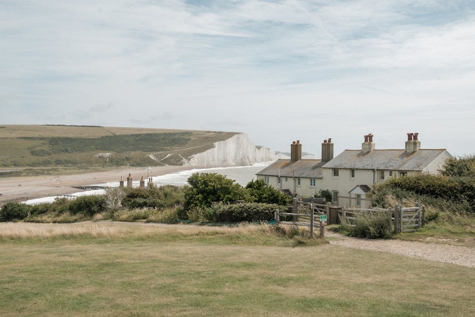 An aerial view of a coastal area showing a sandy beach with gentle waves breaking along the shoreline, bordered by grassy cliffs and open fields. Near the coast, there are several residential houses, some with visible roofs and driveways, situated on a slightly elevated terrain. A narrow pathway runs parallel to the coastline, leading inland through the green landscape. The sky is overcast with soft, diffused lighting, and the overall scene depicts a peaceful seaside community potentially involved in house removals or relocation services, as suggested by the context of home moving logistics. The image aligns with themes of residential move planning, transport of belongings, and local area familiarity, consistent with the services provided by Man with Van Seven Sisters on their page about local moves from Ferry Lane to Bruce Grove.