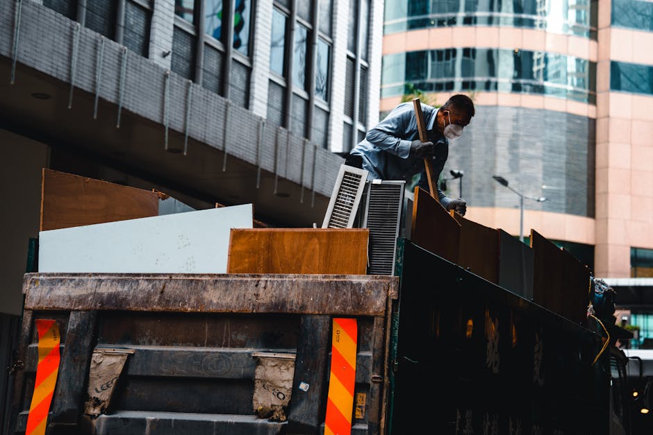 A man wearing a face mask and casual clothing is standing on a flatbed truck loaded with assorted household furniture and waste objects, including wooden cabinets, a white panel, plastic vents, and cardboard packaging materials. He is using a tool to secure or position the items, which are arranged along the edge of the vehicle, preparing for removal or disposal as part of home relocation or furniture transport. The truck is parked in an urban area with modern multi-storey office buildings made of glass and concrete in the background, and the lighting suggests daytime. The scene illustrates the process of loading household items and bulky waste for house removals or clearance services, consistent with professional removals operations by Man with Van Seven Sisters.