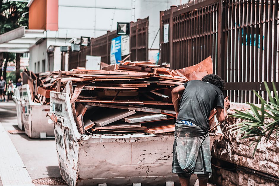 A person with curly hair, wearing a black t-shirt and plaid shorts, stands next to a large, white, open skip filled with discarded roof tiles, wooden planks, and other bulky waste materials. The skip, situated on a paved sidewalk, is positioned against a brown metal fence that runs alongside the street. In the background, there are a few pedestrians, street signs, and a building with orange and white walls. The scene depicts a waste collection or removal process typical during home relocation or clearance, with the materials prepared for transport. This setting aligns with the services provided by Man with Van Seven Sisters under their removals category, focusing on bulky waste removal and local pickup options in Seven Sisters.