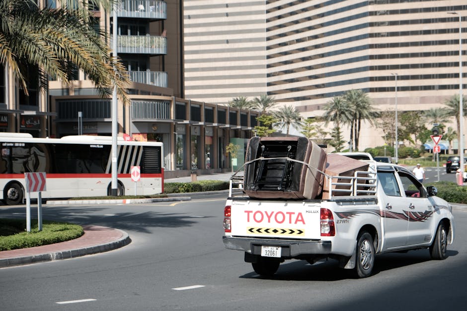 A man wearing a face mask and casual clothing is standing on a flatbed truck loaded with assorted household furniture and waste objects, including wooden cabinets, a white panel, plastic vents, and cardboard packaging materials. He is using a tool to secure or position the items, which are arranged along the edge of the vehicle, preparing for removal or disposal as part of home relocation or furniture transport. The truck is parked in an urban area with modern multi-storey office buildings made of glass and concrete in the background, and the lighting suggests daytime. The scene illustrates the process of loading household items and bulky waste for house removals or clearance services, consistent with professional removals operations by Man with Van Seven Sisters.
