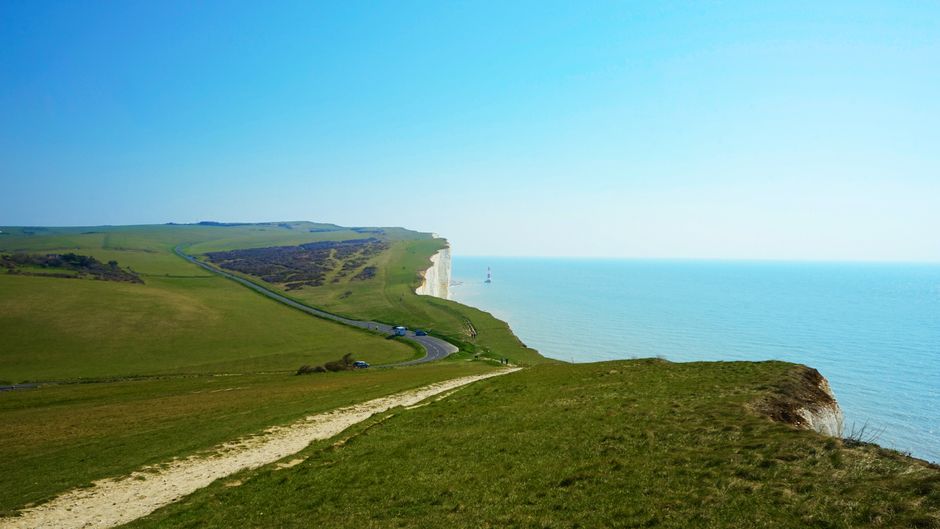 A landscape view of the Seven Sisters cliffs showing a narrow winding road along the green grassy hillside leading towards the white chalk cliffs overlooking the sea. The scene includes a clear blue sky, a few parked cars near the edge of the cliff, and a sailboat visible in the distance on the calm waters of the English Channel. The terrain features a mix of lush grass and patches of exposed chalk, with a footpath in the foreground. This scenic coastal environment exemplifies the natural beauty surrounding the area, which is relevant to the context of house removals and home relocation services offered by Man with Van Seven Sisters, particularly in terms of navigating distinctive coastal landscapes during furniture transport and moving logistics.