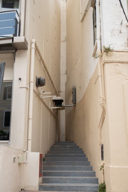 A narrow outdoor staircase located between two pale-colored building walls, leading upwards with a series of dark gray steps made from concrete or stone. On the left side, there are exposed white plastic piping and an outdoor electrical meter box attached to the wall, with some cables and conduits running along the surface. On the right side, a small plant is growing near the base of the wall, which has patches of dirt and minor discoloration. The scene appears to be part of a residential or commercial property in an urban area, with no visible people or moving equipment in the image. This setting reflects typical features of home relocation logistics involving narrow passages, with the exterior environment illuminated by natural daylight. Man with Van Seven Sisters may use such spaces during furniture transport or packing and moving operations within the Seven Sisters area.