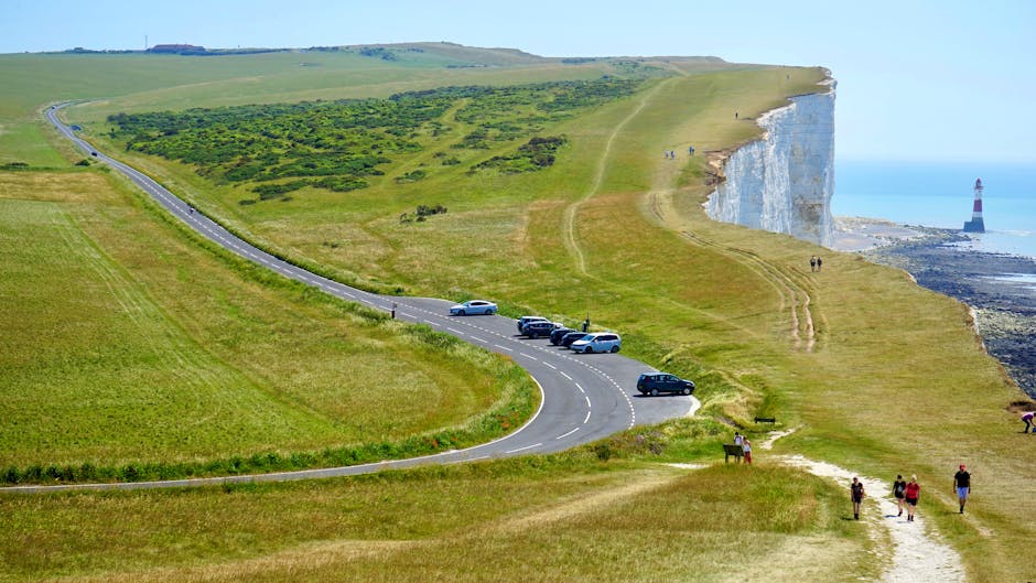A curved coastal road with several parked cars, including a silver sedan and multiple black vehicles, is situated on a grassy hillside overlooking white chalk cliffs and the sea. The road runs through a green landscape with open fields, and leading paths are visible across the terrain. Pedestrians are walking along the grassy paths near the cliff edge, some in groups and others alone. In the background, a black and white lighthouse stands near the shoreline, and the sky is clear with bright, natural lighting enhancing the vibrant colours of the scenery. The image depicts a scenic location suitable for outdoor activities or sightseeing, with the natural environment highlighting the importance of safe transportation and managing mobility in rural and coastal areas, relevant to house removals or home relocation services offered by Man with Van Seven Sisters.