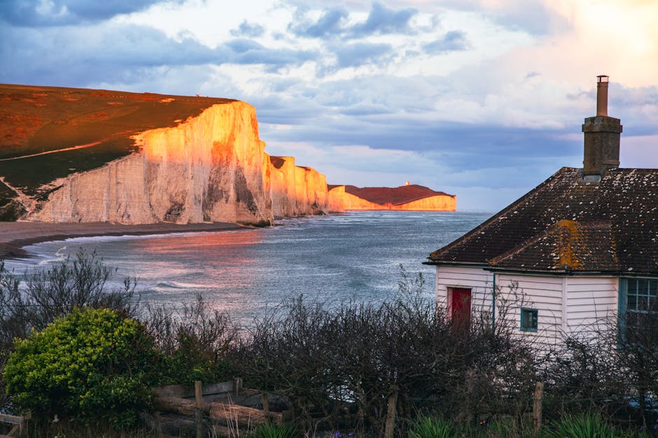 A white, traditional house with a dark, weathered tiled roof and a brick chimney is situated in the foreground, partially obscured by leafless bushes and small green shrubs in a seaside setting. The house is positioned on a slope overlooking a calm sea, with gentle waves lapping against the shoreline. In the background, towering white chalk cliffs are illuminated by the warm glow of the setting sun, casting an orange hue onto their surfaces. The sky above is partly cloudy, with patches of blue and scattered clouds, contributing to the golden hour lighting. This scenic coastal landscape highlights the natural environment located near Haringey, suitable for house removals and home relocation services by Man with Van Seven Sisters, demonstrating the context of transporting furniture and belongings through diverse terrains during a moving process.